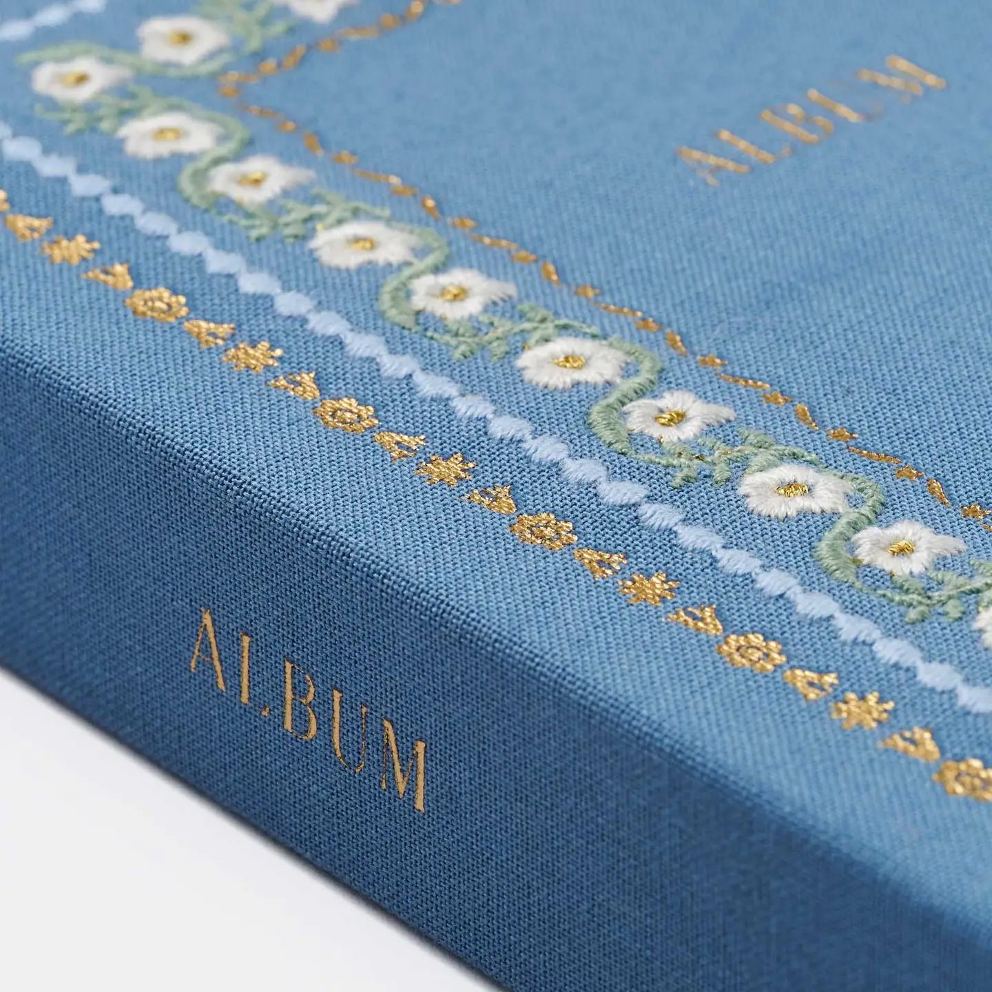 A close-up of a blue fabric-covered album or notebook cover with gold foil lettering reading 'ALBUM' and a decorative border of white flowers and gold details.