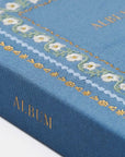 A close-up of a blue fabric-covered album or notebook cover with gold foil lettering reading 'ALBUM' and a decorative border of white flowers and gold details.