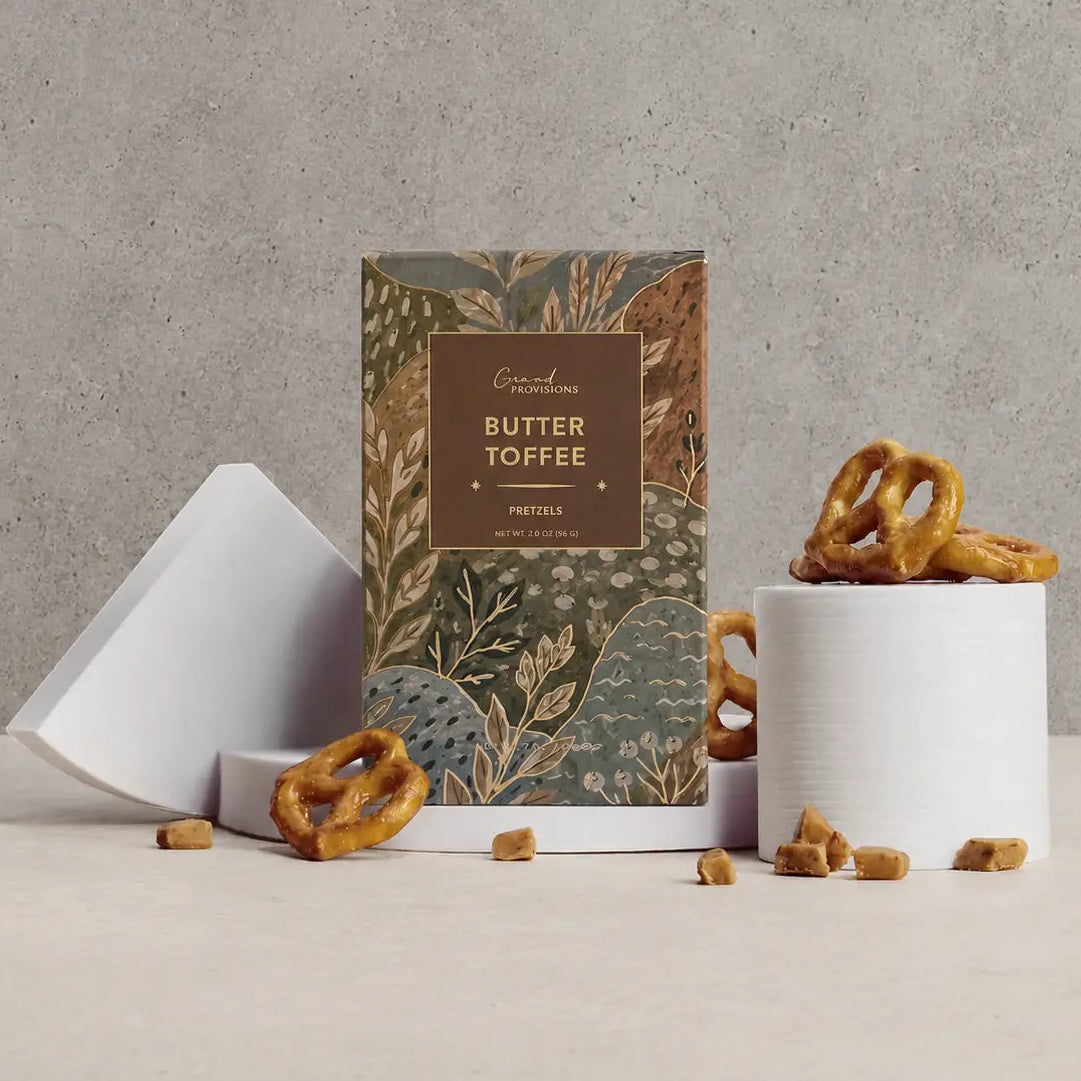 A gourmet butter toffee pretzel box by Goodness Bakery, styled on a light beige surface against a textured gray concrete wall. The packaging features a botanical pattern in muted green, gold, and brown tones.