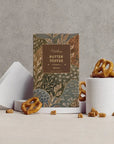 A gourmet butter toffee pretzel box by Goodness Bakery, styled on a light beige surface against a textured gray concrete wall. The packaging features a botanical pattern in muted green, gold, and brown tones.