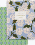 Small notebook with periwinkle background and pink and white hydrangea flowers. The second notebook is green with blue wavy lines and small dots.