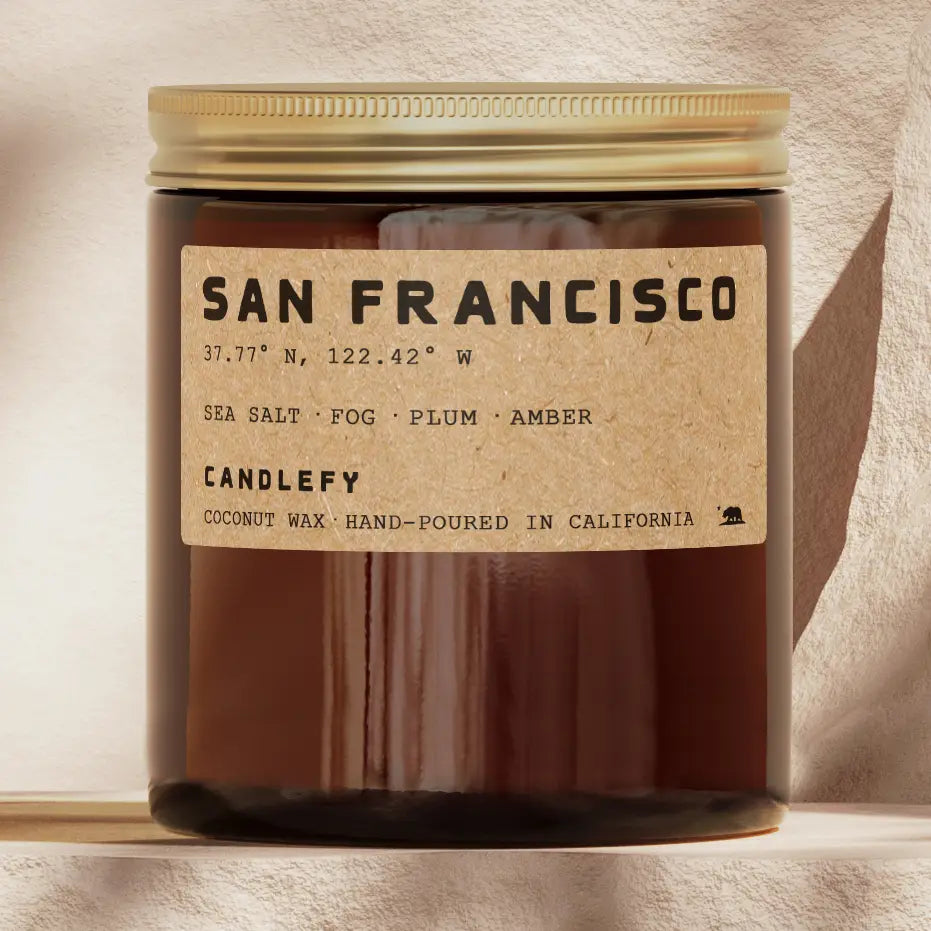 An amber glass jar candle with a gold screw-top lid and a kraft paper label reading 'SAN FRANCISCO' and 'CANDLEFY'. The candle is photographed against a warm beige textured wall with soft shadows.