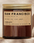 An amber glass jar candle with a gold screw-top lid and a kraft paper label reading 'SAN FRANCISCO' and 'CANDLEFY'. The candle is photographed against a warm beige textured wall with soft shadows.