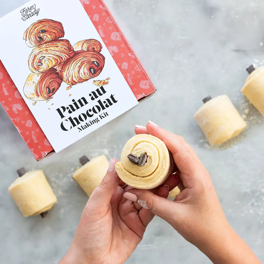 Hands holding a chocolate roll with a 'Pain au Chocolat Making Kit' box in the background.