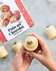 Hands holding a chocolate roll with a 'Pain au Chocolat Making Kit' box in the background.