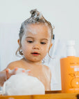 Child with soap bubbles in hair next to a bottle of shampoo