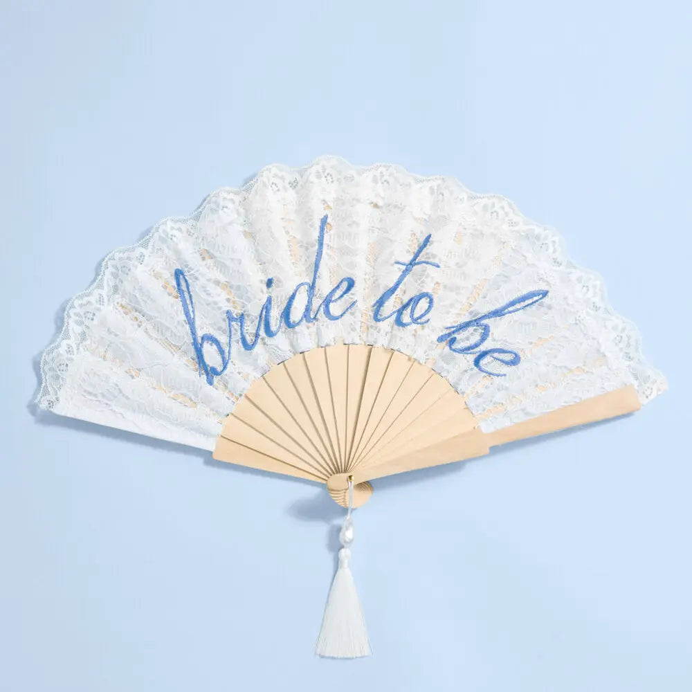 A folding hand fan with a cream lace overlay and blue script reading 'bride to be', attached to a light wood frame with a white tassel, set against a light blue background.