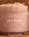 A jar of NCLA Beauty's "Hey, Sugar" exfoliating all-natural body scrub, placed against a warm, brown background. The scrub has a textured, whipped appearance and is surrounded by pieces of cinnamon sticks, emphasizing its natural ingredients and soothing qualities. The jar is labeled with soft, white text detailing the product's name and weight (8.8 oz / 250 g).