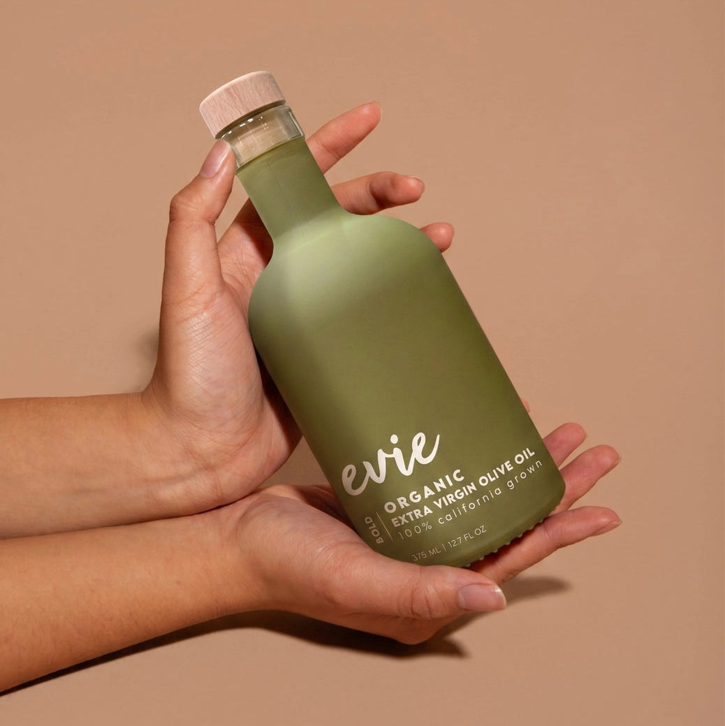 A person's hands holding a frosted olive green bottle of evie Organic Extra Virgin Olive Oil against a solid warm beige background.