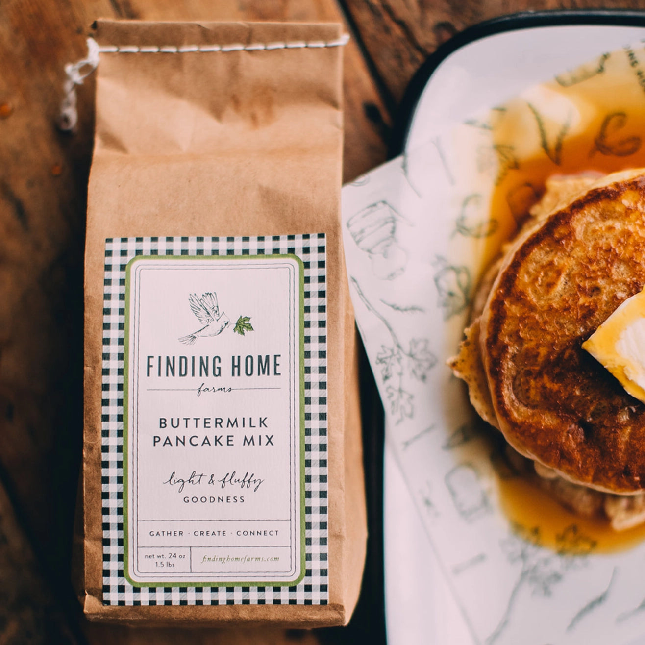 Bag of Finding Home Farms Buttermilk Pancake Mix next to a stack of pancakes with syrup on a wooden surface.