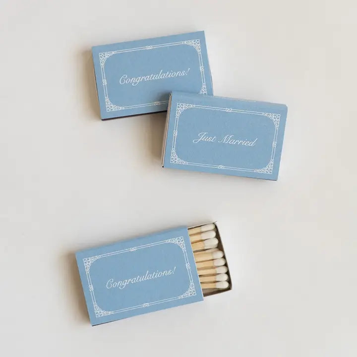 Three light blue matchboxes with white decorative borders and script text reading Congratulations, Just Married, and Congratulations, arranged on a plain light background. One matchbox is open and shows white matches inside.