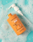 An orange bottle of dabble & dolly Tangerine Gel bath bomb with a white pump dispenser, placed on a light blue background covered in white foam bubbles.