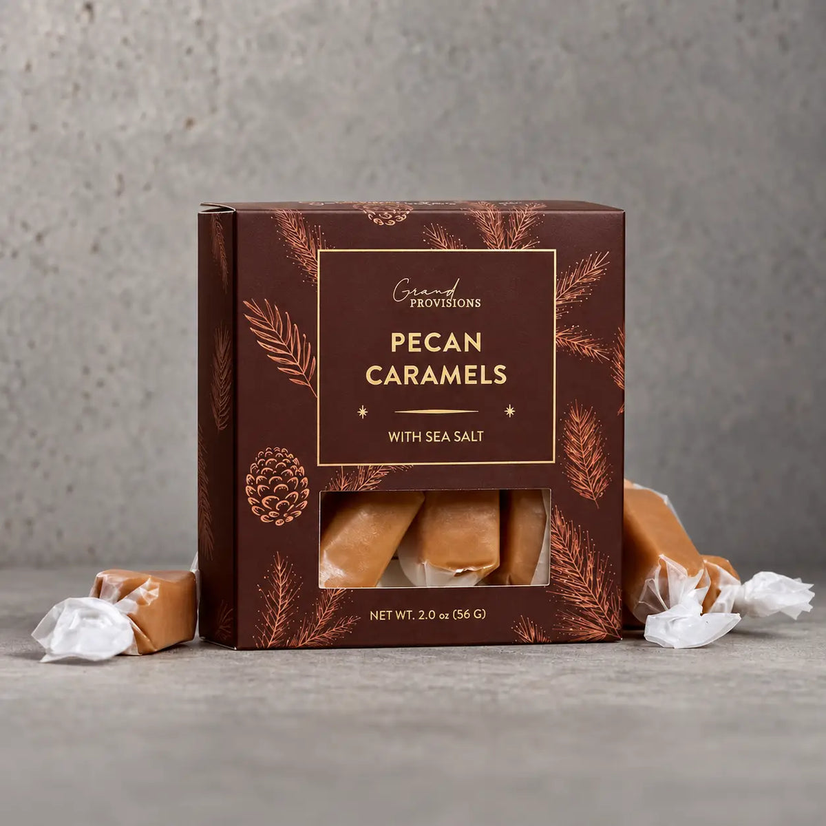 A box of Grand Provisions Pecan Caramels with Sea Salt, displayed on a gray concrete surface with unwrapped caramel candies placed around the box.