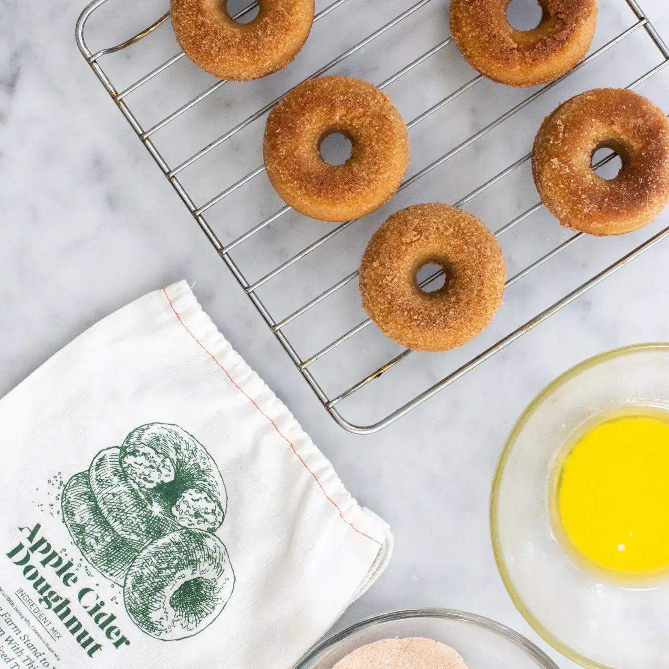 Six baked apple cider doughnuts rest on a metal cooling rack over a light marble surface. A white kitchen towel printed with green apple cider doughnut graphics and text lies nearby, with a glass bowl containing yellow liquid and another bowl partially visible at the bottom right.
