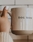 Hand holding a mug with 'DOG MOM' text against a neutral background