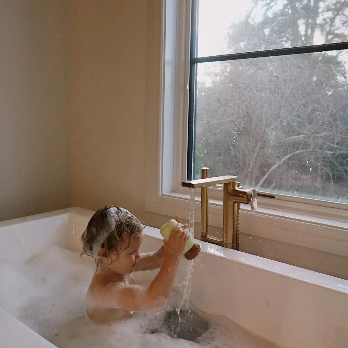Child in a bathtub playing with water near a window