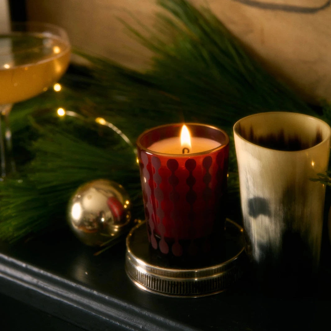 This image shows a cozy, festive scene featuring a lit candle in a red, patterned glass holder. Beside it is another candle or holder in a beige and black gradient pattern. The setting includes holiday greenery, a small string of warm lights, and a reflective silver ornament. In the background, there’s a cocktail glass, adding to the warm, elegant holiday atmosphere.






