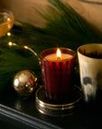 This image shows a cozy, festive scene featuring a lit candle in a red, patterned glass holder. Beside it is another candle or holder in a beige and black gradient pattern. The setting includes holiday greenery, a small string of warm lights, and a reflective silver ornament. In the background, there’s a cocktail glass, adding to the warm, elegant holiday atmosphere.






