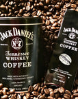 A black metal tin and matching black pouch of Jack Daniel's Tennessee Whiskey Coffee surrounded by scattered roasted coffee beans.