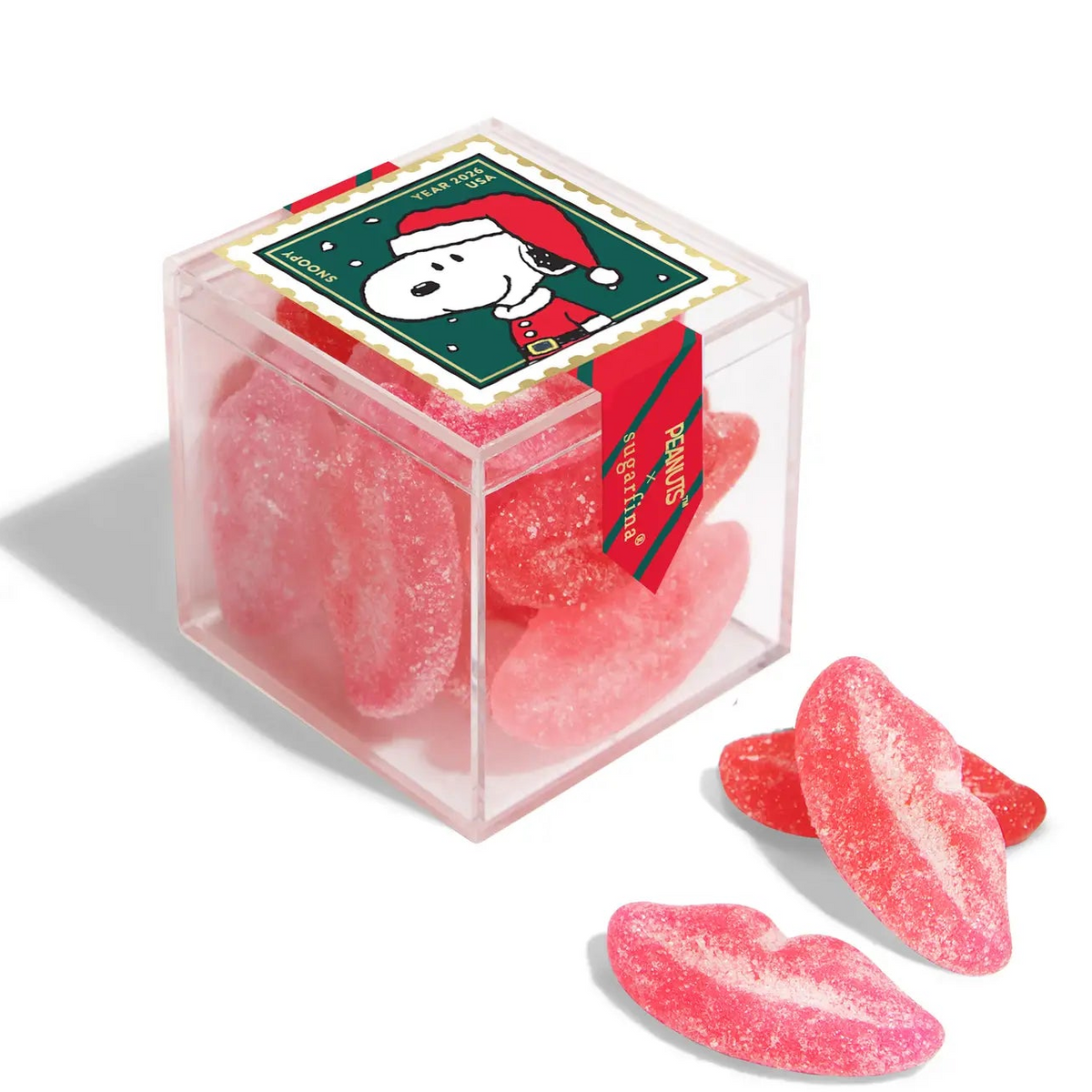 A clear square gift box filled with pink sugar-coated gummy candies, topped with a holiday-themed label featuring Snoopy in a Santa hat. Three lip-shaped gummies are placed in front on a white background.