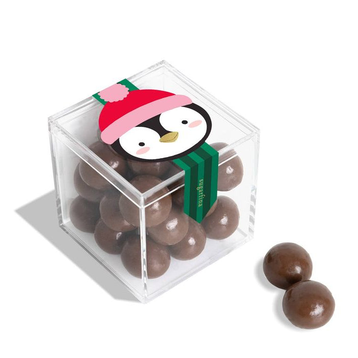 A clear acrylic cube container filled with chocolate-covered round candies, with a penguin illustration on the lid and two additional candies placed beside it on a white background.