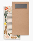 A kraft paper shopping list notepad with a dark gray label holder on the front, partially covering the back cover which features a cream-colored background with hand-drawn illustrations of vegetables, bread, eggs, and a milk bottle.