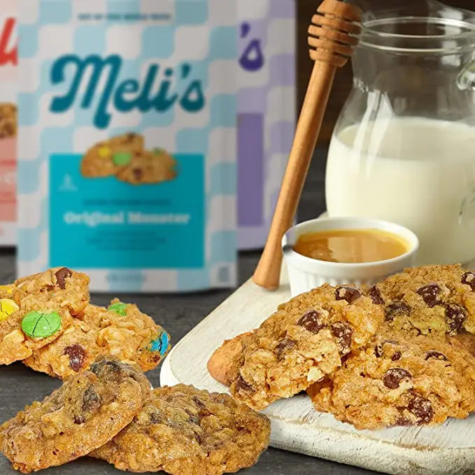 Meli's Original Monster cookies displayed on a white cutting board with a glass jar of milk, a wooden honey dipper, and a small cup of honey, with the product packaging in the background.