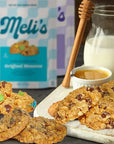 Meli's Original Monster cookies displayed on a white cutting board with a glass jar of milk, a wooden honey dipper, and a small cup of honey, with the product packaging in the background.