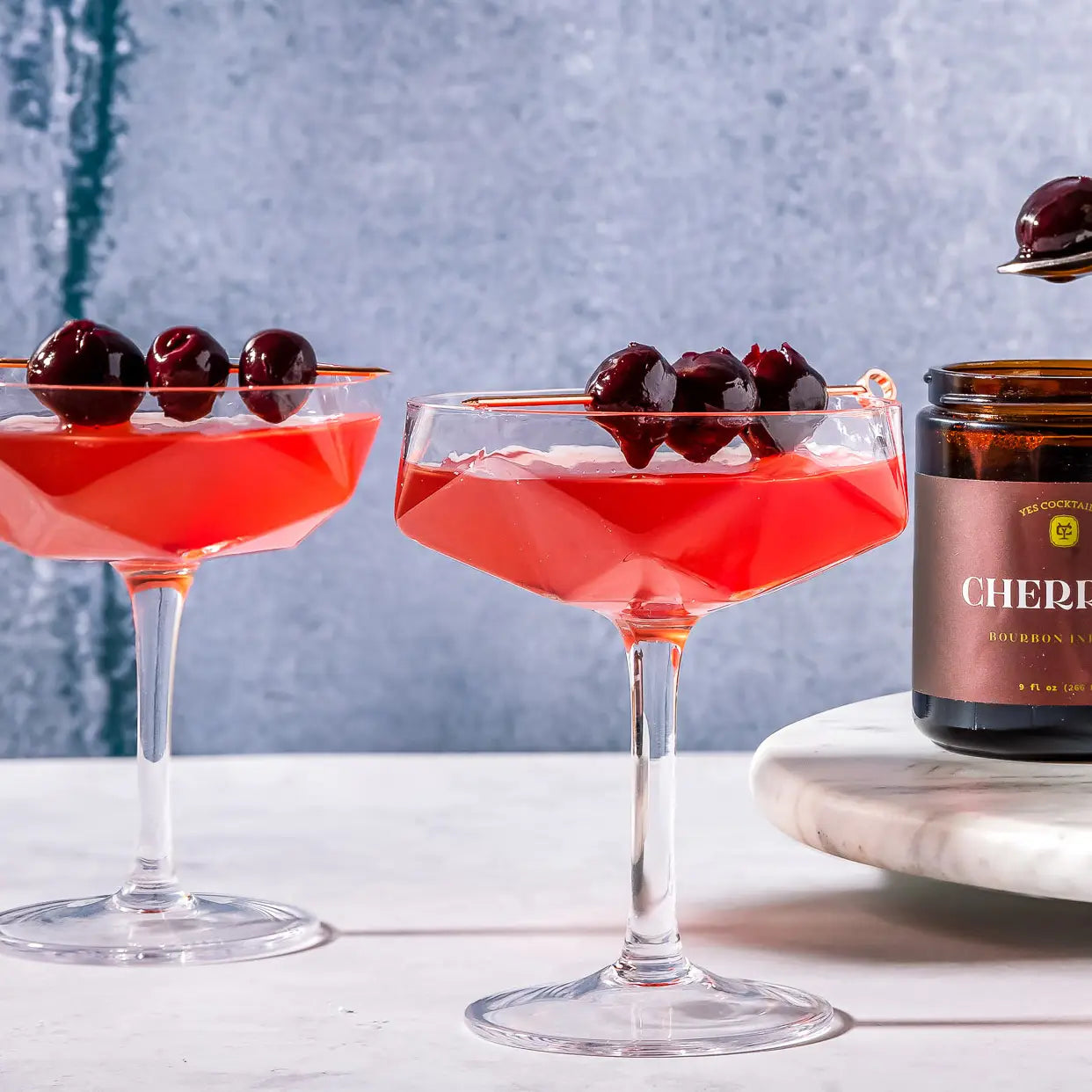 Two red cocktails with cherries in glass coupes on a white surface, with a jar of cherries in the background.