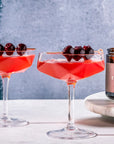 Two red cocktails with cherries in glass coupes on a white surface, with a jar of cherries in the background.