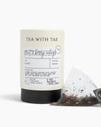 A tea product shot on a clean white background, featuring a cylindrical black canister with a cream label reading 'TEA WITH TAE' and a clear teabag with loose tea leaves and a black brand tag in front.
