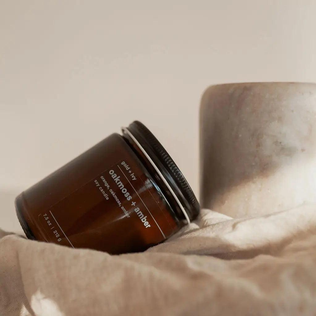 Brown candle jar labeled 'Oakmoss & Amber' on a textured surface with a neutral background