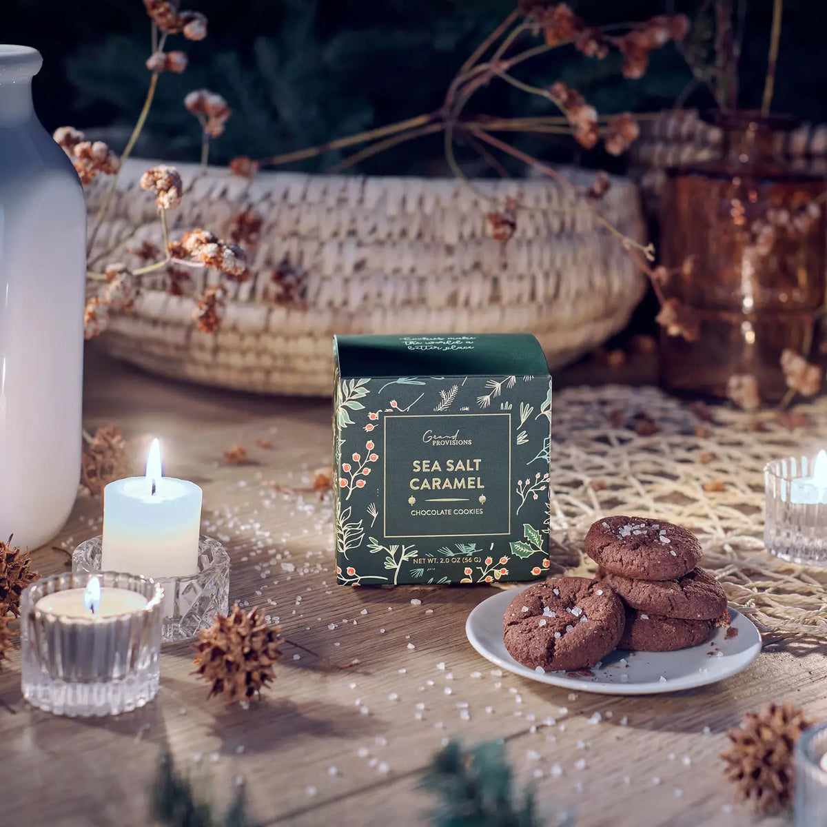 A boxed gourmet chocolate cookie called 'Sea Salt Caramel' sits on a rustic wooden table styled with lit candles, dried flowers, pinecones, and a plate of chocolate cookies. The box is dark green with botanical illustrations.