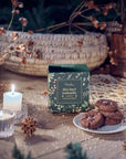 A boxed gourmet chocolate cookie called 'Sea Salt Caramel' sits on a rustic wooden table styled with lit candles, dried flowers, pinecones, and a plate of chocolate cookies. The box is dark green with botanical illustrations.
