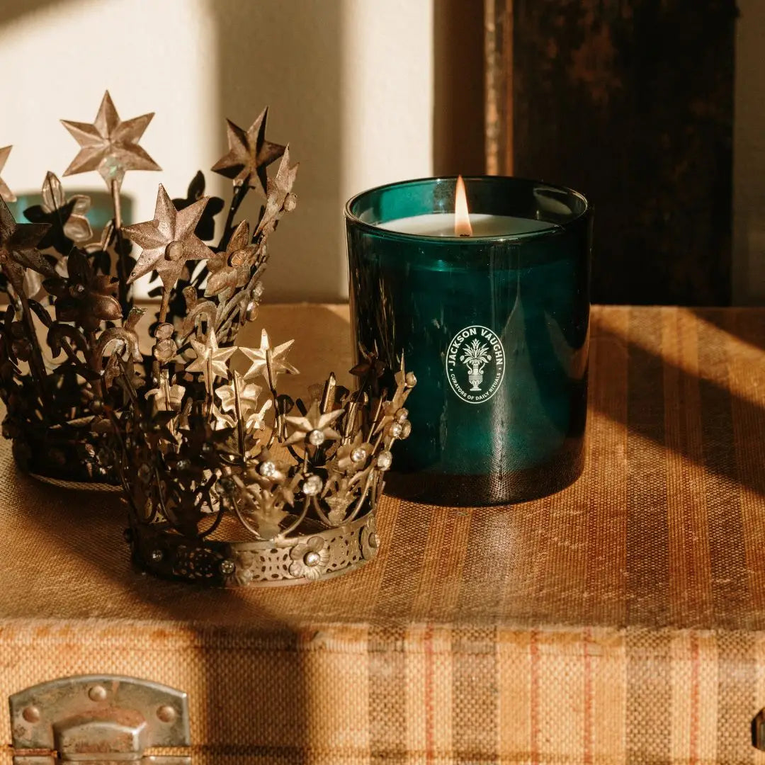 A dark teal glass candle with a white wax fill and a visible flame sits on a rustic wooden surface next to two ornate metallic star crowns. The candle has a circular white logo on the front.