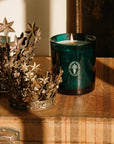 A dark teal glass candle with a white wax fill and a visible flame sits on a rustic wooden surface next to two ornate metallic star crowns. The candle has a circular white logo on the front.
