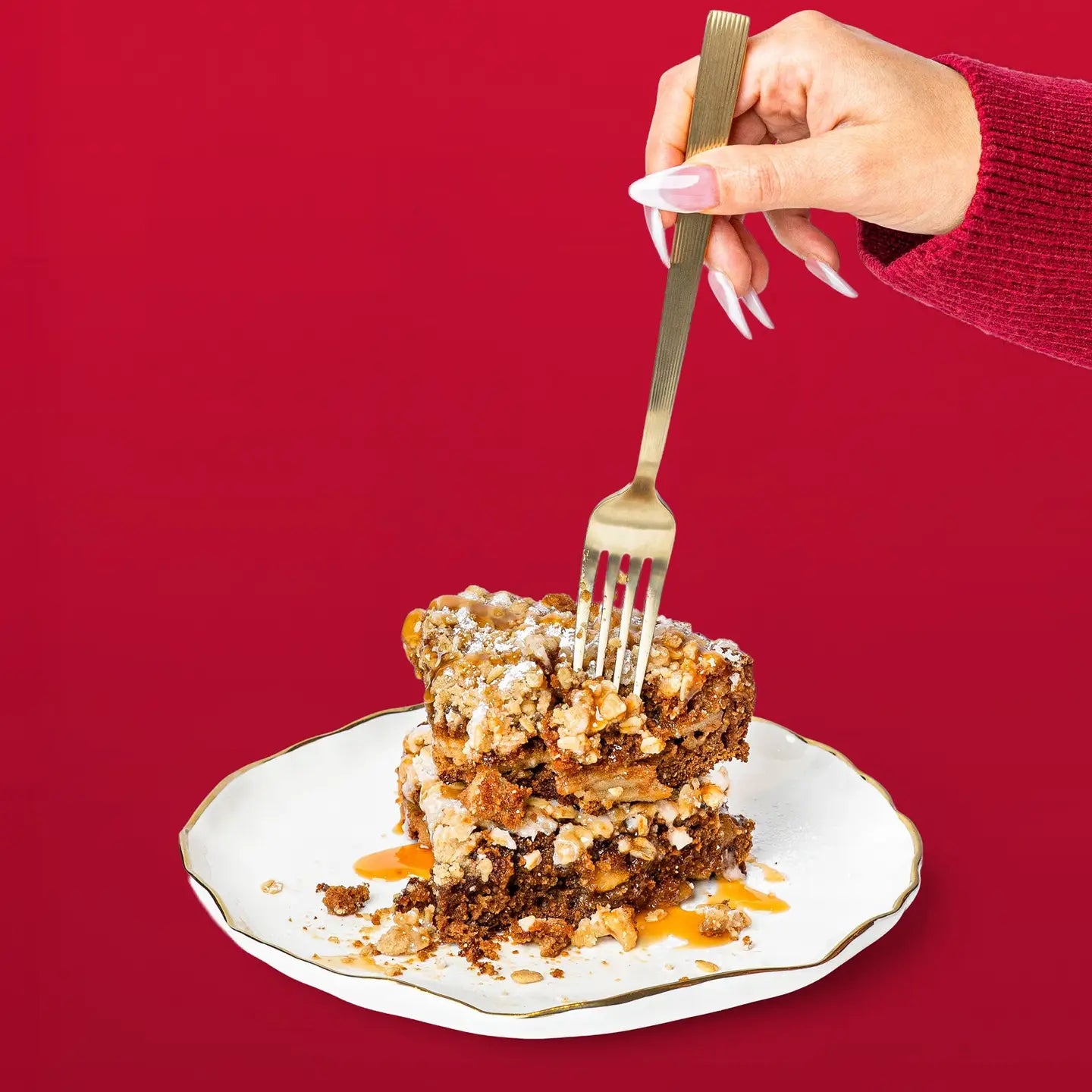 A hand with long pink nails holds a gold fork over a slice of layered dessert on a white plate with a gold rim, set against a solid red background.