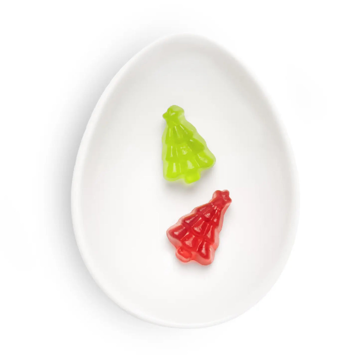 Two small Christmas tree-shaped gummy candies, one green and one red, displayed inside a white ceramic egg-shaped dish on a white background.
