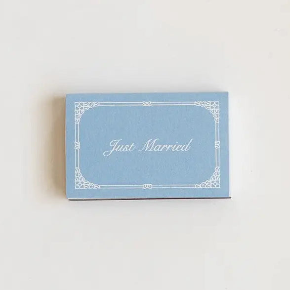 A small rectangular light blue card with a white decorative border and the words 'Just Married' in white script, placed on a plain off-white background.