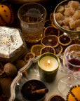 A cozy holiday spread featuring a wooden bowl filled with walnuts, an orange studded with cloves, and a decorative glass holding a drink. The arrangement includes a silver tray with lit candles, dried citrus slices, mulled wine with cinnamon, and a bowl of sugared treats, creating a warm festive atmosphere.