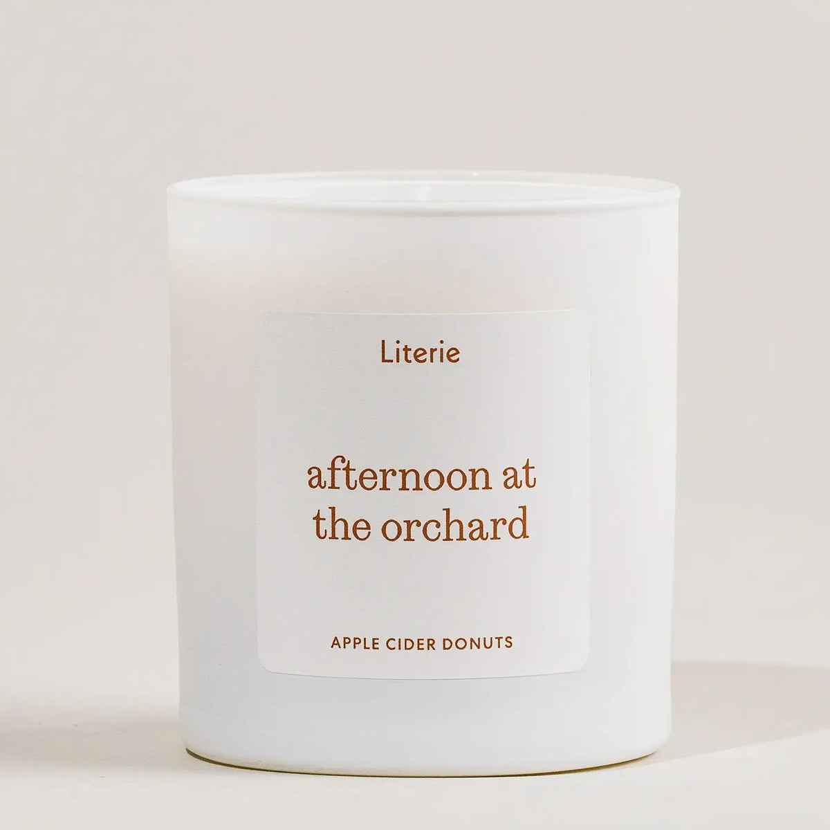 A white glass jar candle with a minimalist white label featuring the scent name 'afternoon at the orchard' in a warm brown font, with 'Literie' above and 'APPLE CIDER DONUTS' below, set against a clean off-white background.