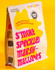 A package of Mojave Mallows S'more Speckled Marshmallows displayed against a bright yellow background. The packaging features bold magenta text, highlighting chocolate chip marshmallows dusted with graham crackers, ideal for snacking or toasting.