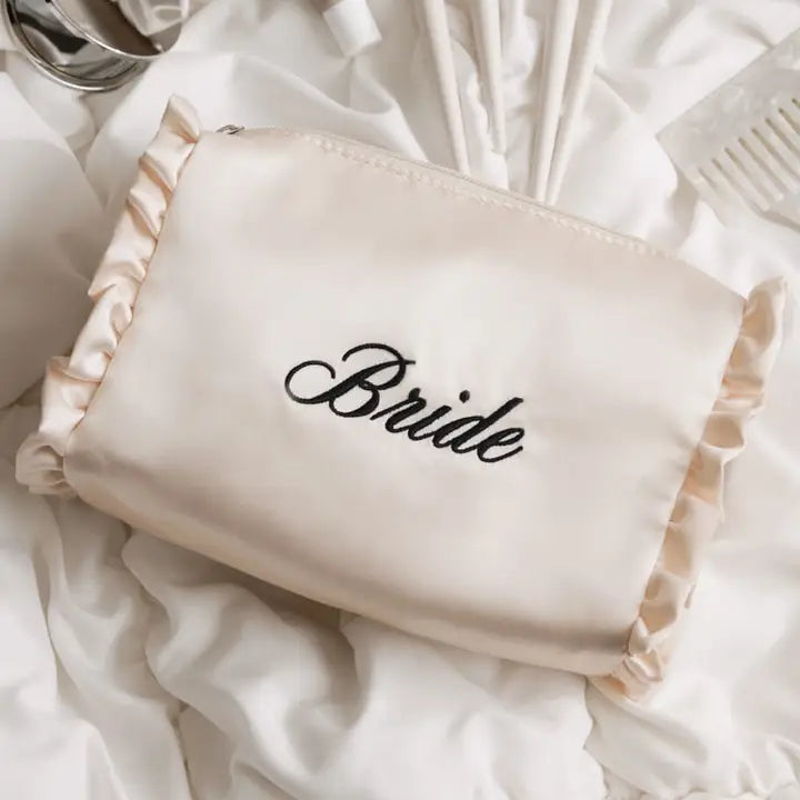 A cream satin bridal makeup bag with ruffled trim and the word 'Bride' embroidered in black script, displayed on a bed of white satin sheets.