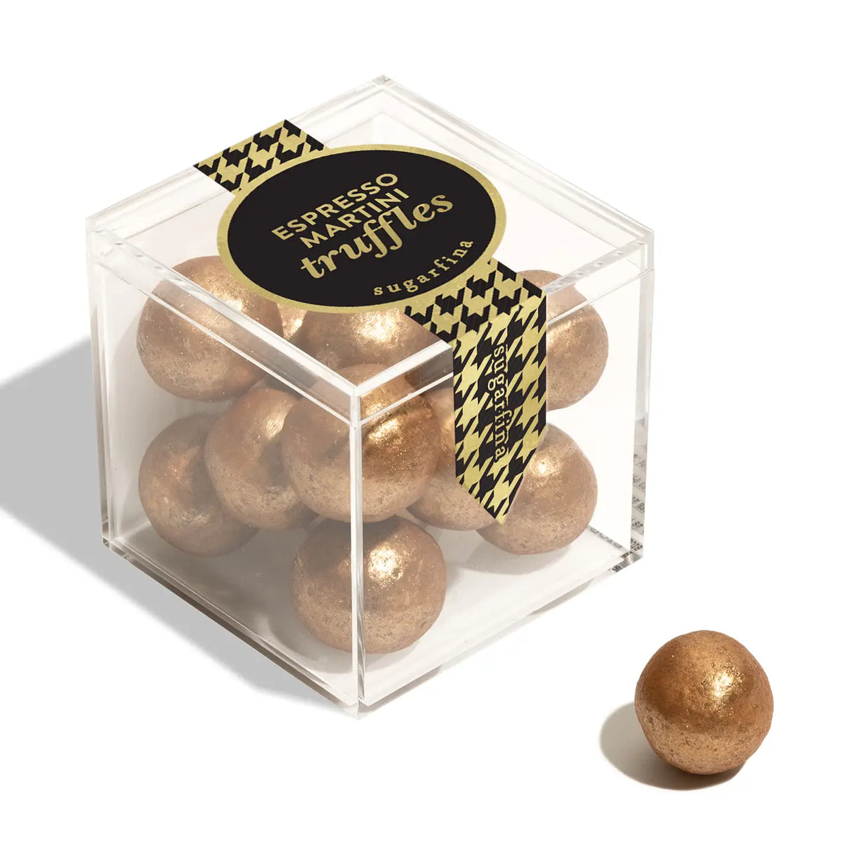 A clear acrylic cube box filled with gold-glittered espresso martini truffles, with a black and gold houndstooth ribbon label. A single truffle sits outside the box on a clean white background.