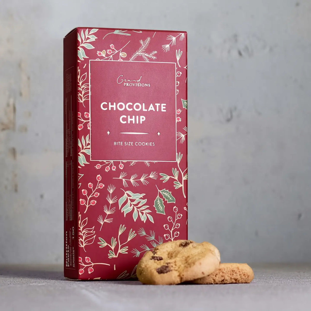 A red box of Grand Provisions Chocolate Chip bite size cookies with botanical illustrations, standing on a gray surface with two cookies in front.
