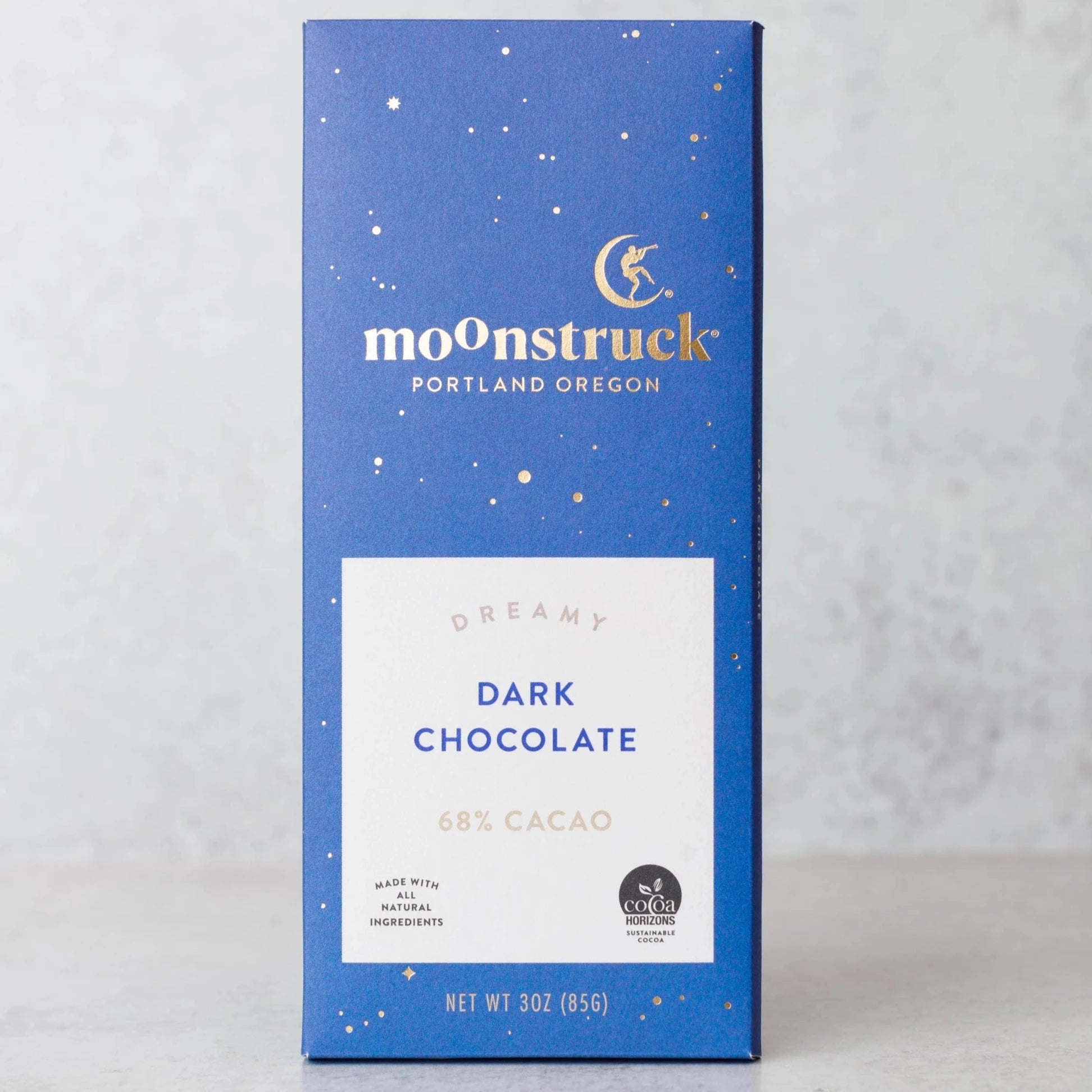 A blue box of Moonstruck Dark Chocolate, labeled as 'Dreamy Dark Chocolate 68% Cacao.' The box design includes white and gold star-like specks, with the text 'Portland Oregon' beneath the Moonstruck logo. The label notes that the chocolate is made with all-natural ingredients and carries the Cocoa Horizons Foundation logo
