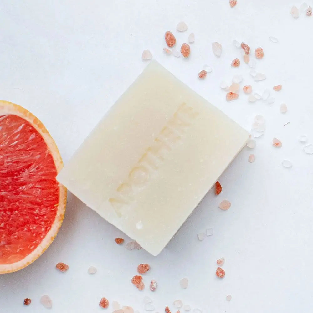 A rectangular bar of pale cream soap with the brand name 'ANOTHERE' embossed on it, styled on a white surface with scattered pink Himalayan salt crystals and a sliced grapefruit half visible on the left.