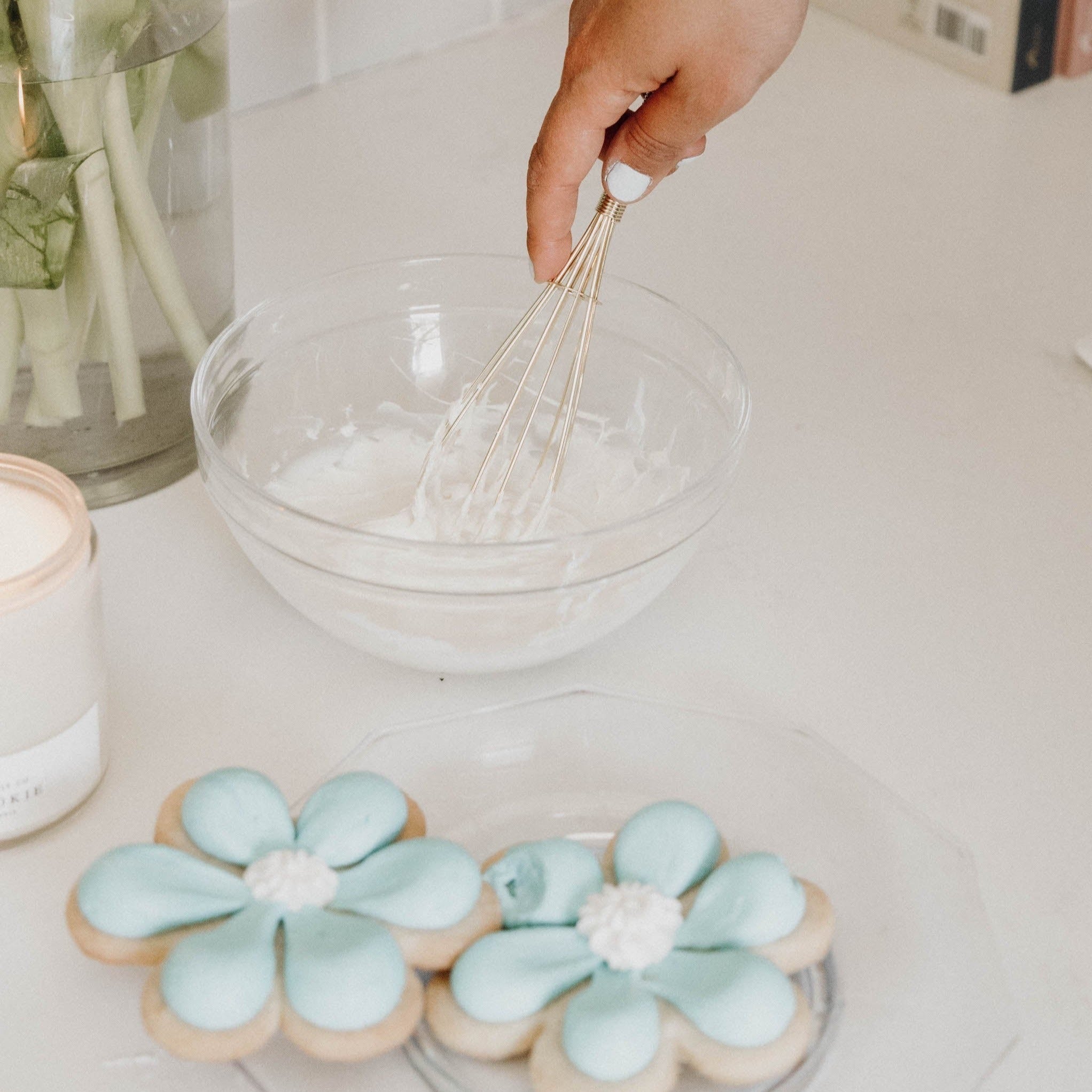 Gold whisk mixing frosting in bowl next to plate of blue flower sugar cookies.