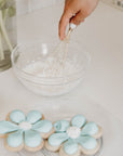 Gold whisk mixing frosting in bowl next to plate of blue flower sugar cookies.