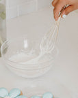 Gold whisk mixing frosting in a glass bowl